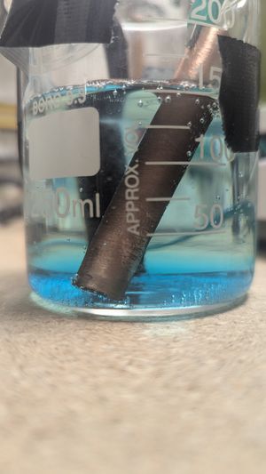 A small 250mL beaker with two copper tubes acting as anode and cathode showing bubbles and a faint blue hue in the solution