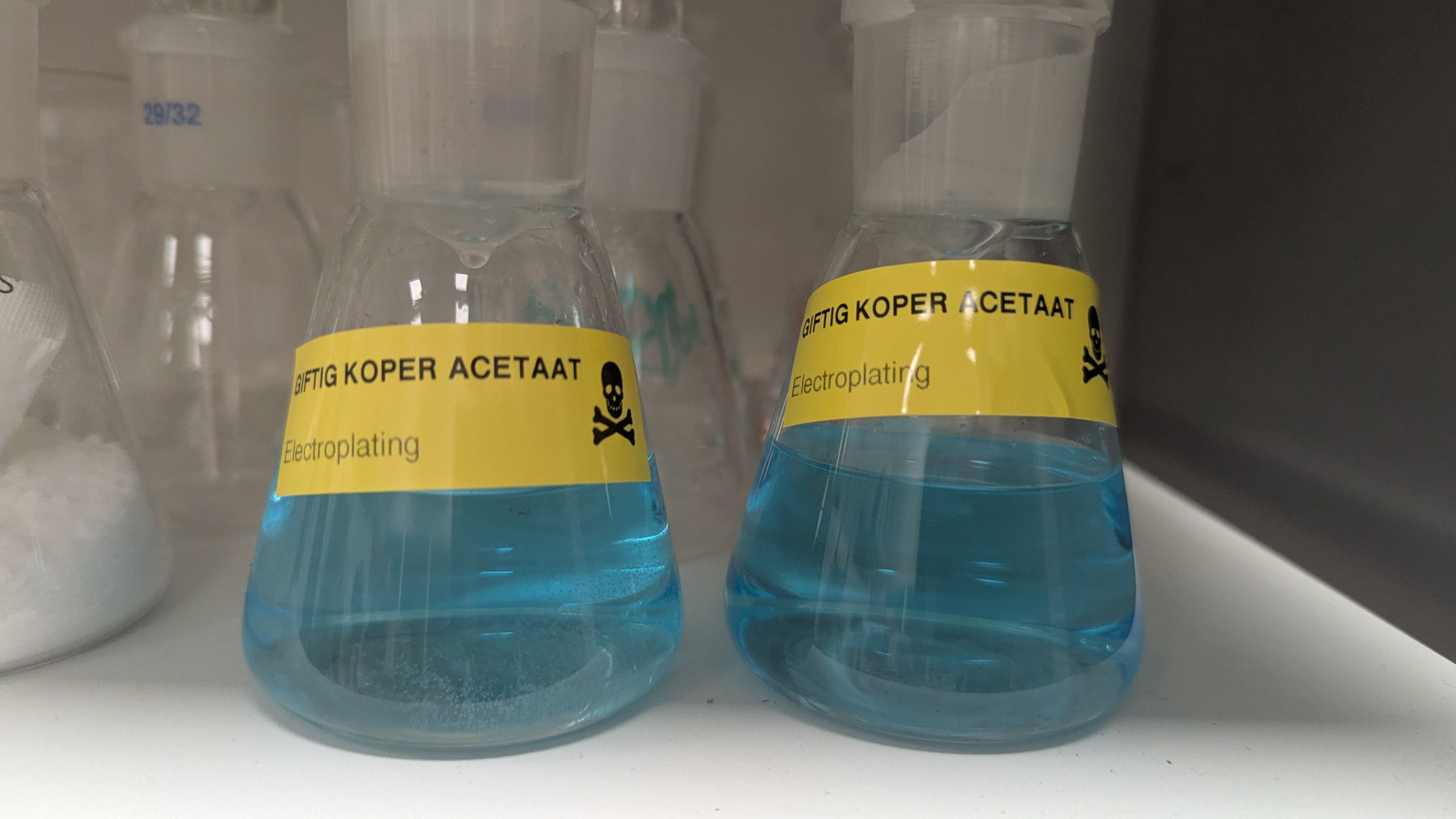 Two 125mL erlenmeyers with the blue copper acetate solution and a sticker with a skull saying in dutch "Posion: Copper Acetate"