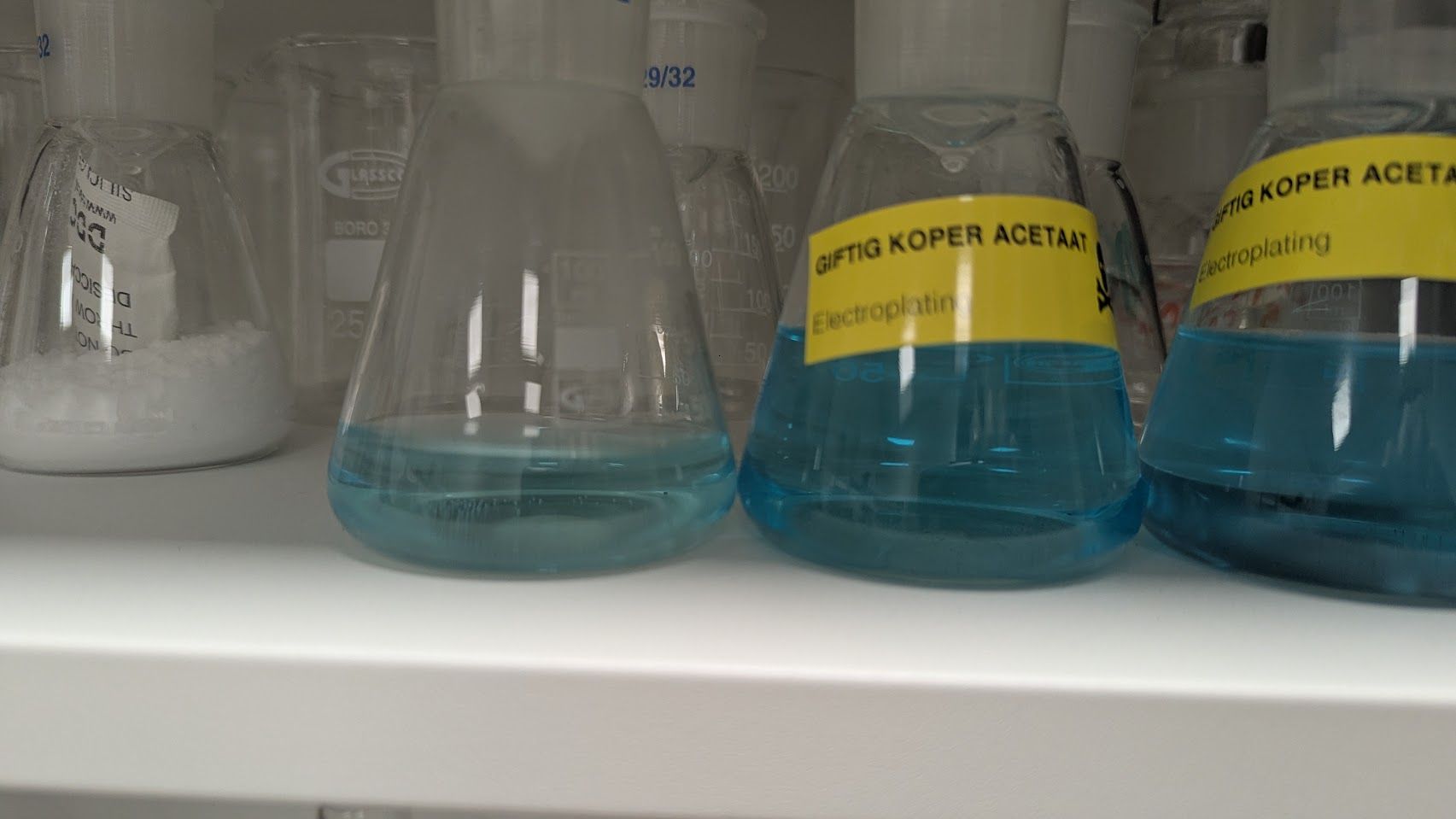 A 125mL erlenmeyer with transparent blue copper acetate solution from the hydrogen peroxide process, next to the two previous erlenmeyers